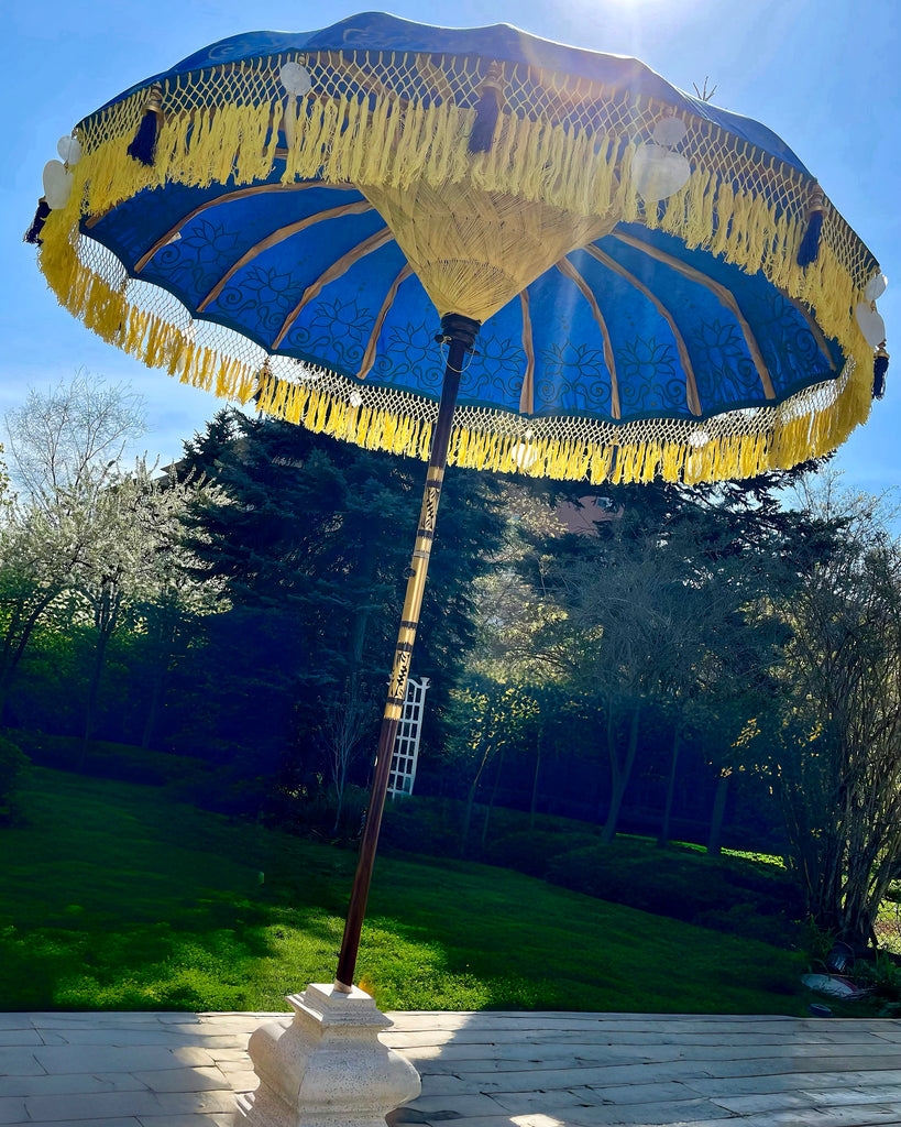 A large blue and yellow Baliaric Parasol Carina Parasol with fringed edges stands open on a sunny day in a garden. The sun glows brightly in the background, casting shadows on the ground. Lush green trees and grass surround this stunning piece of outdoor decor.