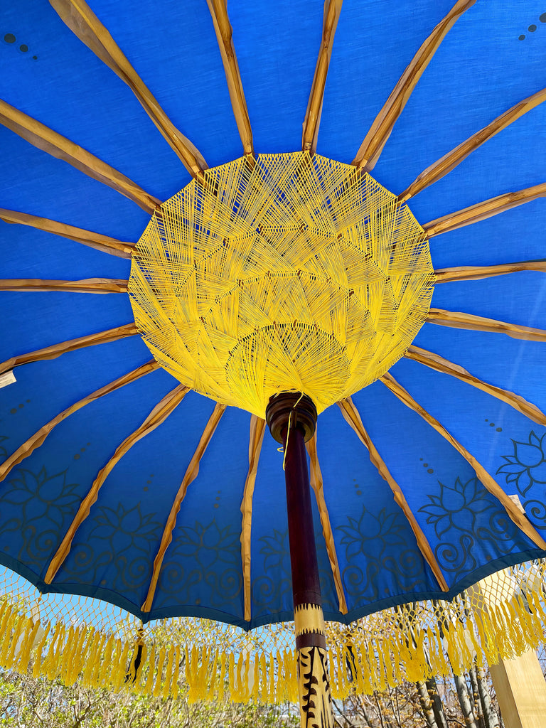 The Baliaric Parasol's Carina Parasol, featuring a blue canopy adorned with exquisite yellow fringe patterns, brings an elegant charm to any outdoor setting. This artisanal parasol boasts radial yellow supports and a central wooden pole, beautifully complementing the natural scenery of trees and blue skies.