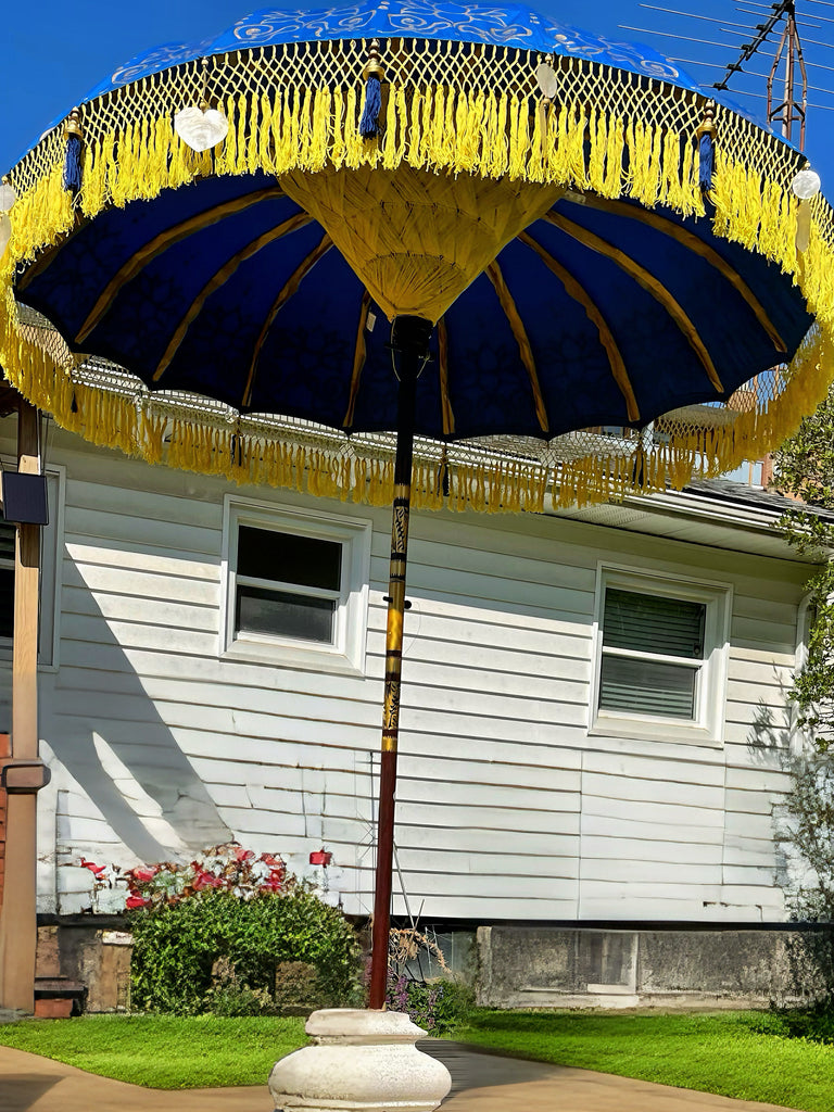 A large and ornate Baliaric Parasol's Carina Parasol, featuring a blue and yellow fringe canopy, stands in a garden before a white house. The handmade parasol's base sits on the green lawn, surrounded by flowers and shrubs, enhancing the outdoor decor.