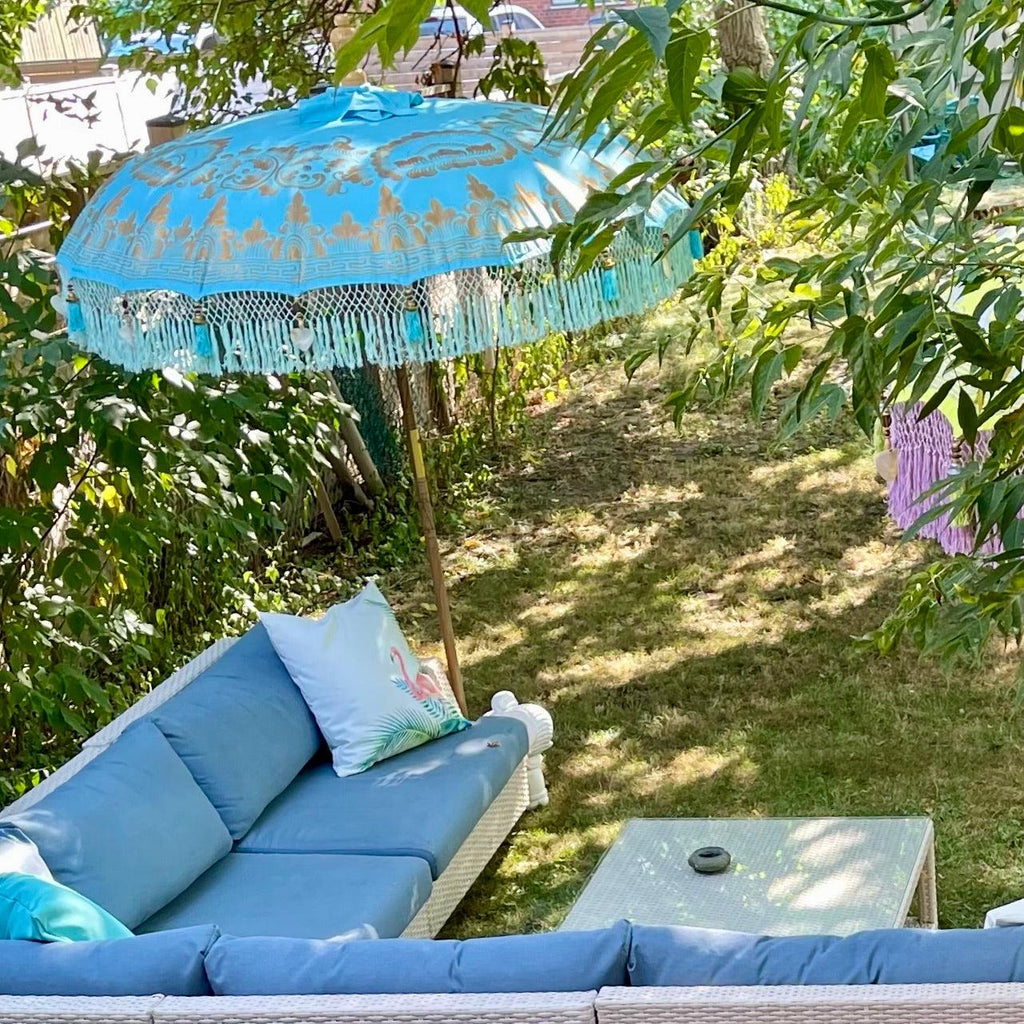 In a backyard setting, a blue cushioned outdoor sofa complements a decorative Big Jack Parasol by Baliaric Parasol that provides shade. Nearby is a small glass table featuring an ashtray, all surrounded by lush greenery and trees that enhance the charm of the outdoor decor.