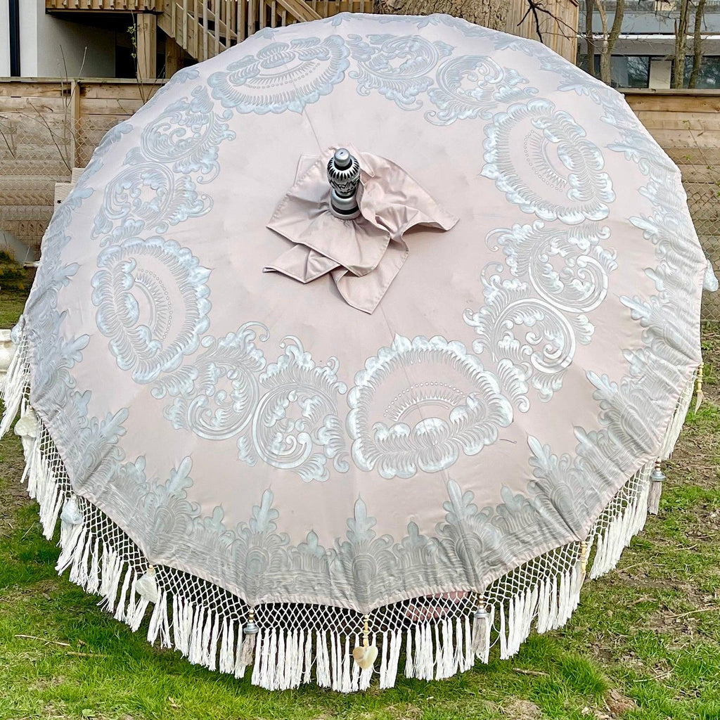 The Baliaric Parasol's Sandy Parasol showcases tropical elegance, featuring intricate white lace patterns and a fringe trim, open on a grassy area and capturing the essence of Bali. The setting suggests it might be in a yard or garden, with wooden fences visible in the background.