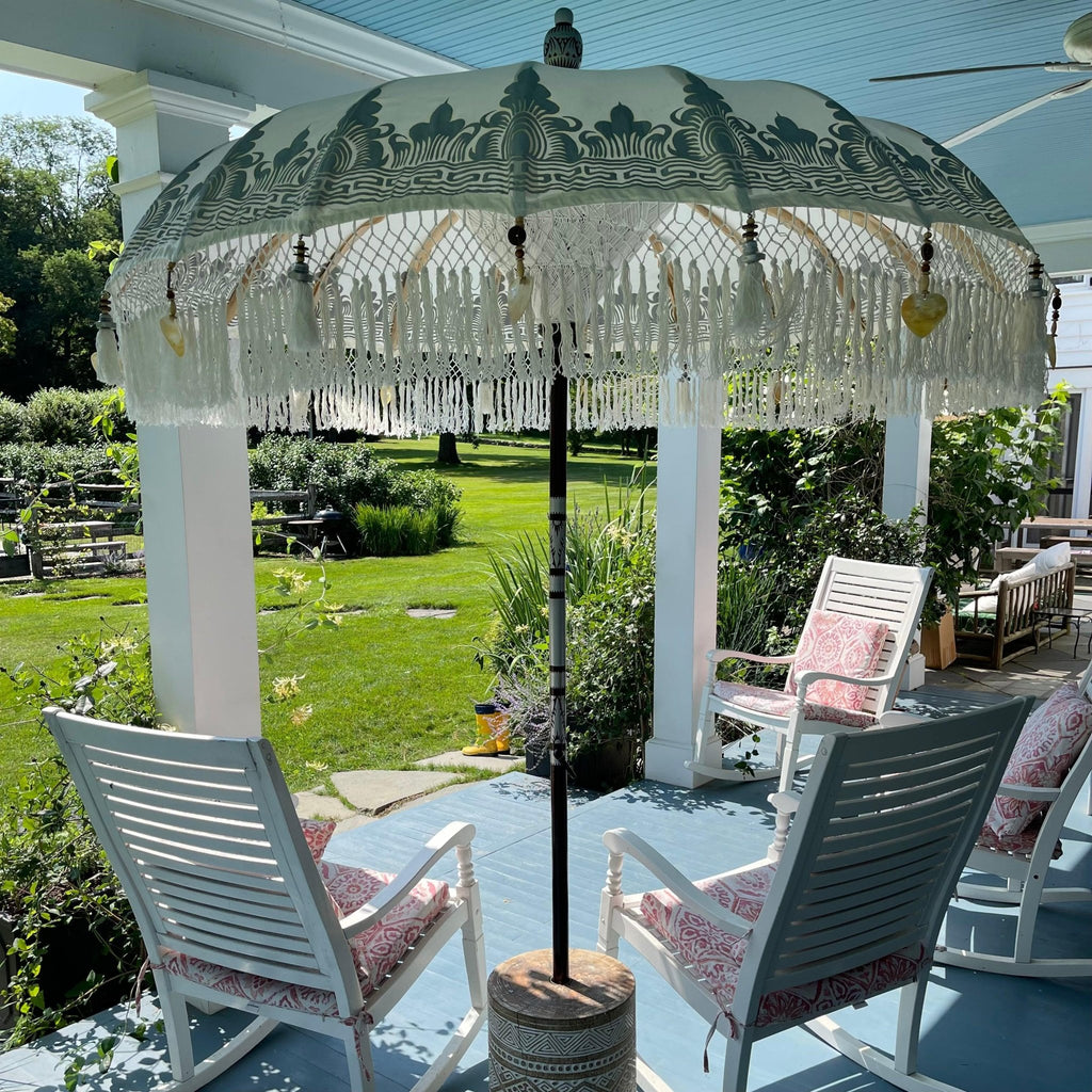 On a sunny porch, two white rocking chairs with pink patterned cushions are positioned under a Baliaric Parasol's White Cloud Parasol, crafted from hand-carved durian wood. This delightful outdoor space offers views of a lush green lawn and garden.