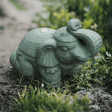 A small Green Elephant Parasol Stand by Baliaric Parasol is nestled among grass and greenery, capturing an exotic elegance in this serene and natural setting.